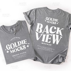 以下が含まれることがあります： 白文字のグレーのTシャツ2枚。1枚のシャツは折りたたまれ、「GOLDIE MOCKS」と「MOCKUP STUDIO」が表示されています。もう1枚のシャツには「BACK VIEW MOCKUP」と表示されています。シルバーのチェーンネックレスが折りたたまれたシャツの上にあります。