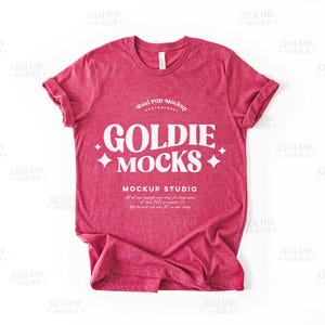 Puede incluir: Una maqueta de camiseta de color rojo jaspeado con el texto "Goldie Mocks" y "Mockup Studio" impreso en la parte delantera. La camiseta está colocada sobre una superficie blanca.