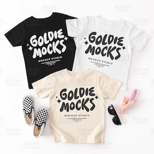 May include: Three t-shirts with the text "Goldie Mocks Mockup Studio" in black lettering on a white background. The shirts are laid out on a white surface with a pair of black and white checkered Vans shoes and some colorful chalk.