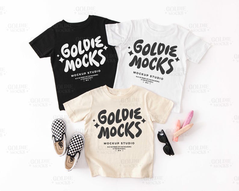 K&ouml;nnte beinhalten: Drei T-Shirts mit dem Text "Goldie Mocks Mockup Studio" in schwarzer Schrift auf wei&szlig;em Hintergrund. Die Shirts liegen auf einer wei&szlig;en Oberfl&auml;che mit einem Paar schwarz-wei&szlig; karierter Vans-Schuhe und etwas buntem Kreide.