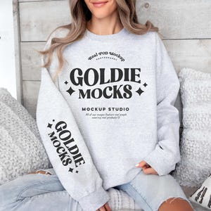 Puede incluir: Una sudadera gris con el texto "Real POD Mockup Photography Goldie Mocks" y "Mockup Studio" en la parte delantera. Las mangas tienen el texto "Goldie Mocks" impreso en ellas.
