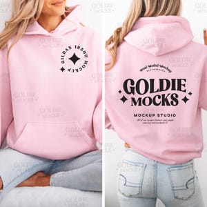 Könnte beinhalten: Ein rosa Hoodie mit einem schwarzen Logo, auf dem "Goldie Mocks" und "Mockup Studio" steht. Das Logo enthält auch eine kleine Diamantenform auf jeder Seite des Textes. Der Hoodie wird von einer Person mit einer blauen Jeans getragen.
