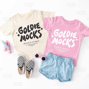 May include: Two children's t-shirts with the text "Goldie Mocks Mockup Studio" printed on them. One shirt is beige with black text and the other is pink with white text.