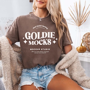 Könnte beinhalten: Ein braunes T-Shirt mit dem Text "GOLDIE MOCKS" in Weiß. Das Shirt wird von einem Model mit blonden Haaren und einem weißen Cardigan getragen. Der Text unter dem Logo lautet "MOCKUP STUDIO" und "All of our images feature real people wearing real products."