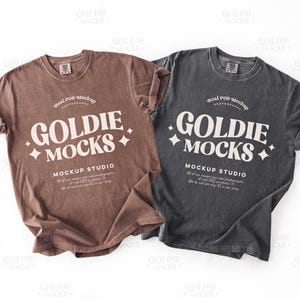 Puede incluir: Se muestran dos camisetas de manga corta, una marrón y otra gris antracita. Cada camiseta presenta el texto "GOLDIE MOCKS" en una fuente estilizada, junto con texto adicional. Las camisetas se presentan sobre un fondo blanco.