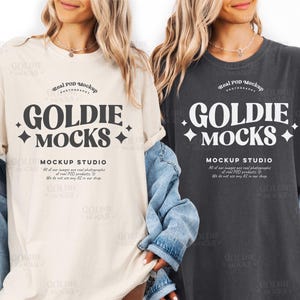 Può includere: Due magliette, una color crema e una grigio antracite, ciascuna con la scritta "GOLDIE MOCKS" in un carattere audace e stilizzato. Il testo è centrato, con le parole "MOCKS" in un corpo più grande. Le magliette presentano anche testo aggiuntivo.