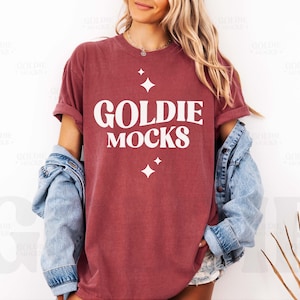 Peut inclure: Un t-shirt marron avec un motif blanc qui dit "Goldie Mocks" et quelques petites étoiles blanches.