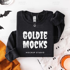 May include: A black crewneck sweatshirt with the text "GOLDIE MOCKS" in white. A silver necklace with a sun pendant is on the shirt. The words "MOCKUP STUDIO" are below the text. Halloween decorations are in the background.