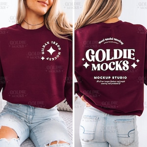 May include: A maroon sweatshirt with a white graphic on the front that says "Gildan 18500" and a white graphic on the back that says "Goldie Mocks" with a white star on each side. The sweatshirt is being modeled by a person wearing blue jeans.