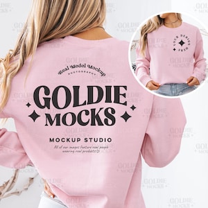 以下が含まれることがあります： 長袖でクルーネックのピンク色のスウェットシャツ。スウェットシャツの背面には、ダイヤモンド型の模様が両側に付いた黒い文字で「GOLDIE MOCKS」と書いてあります。テキストの下には、「MOCKUP STUDIO」というフレーズと、「All of our images feature real people wearing real products.」というテキストがより小さなフォントで書かれています。