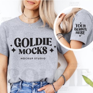 May include: A gray t-shirt mockup with the text "GOLDIE MOCKS MOCKUP STUDIO" printed on the front. The shirt is being worn by a person with long blonde hair. The image also shows a close-up of the shirt's sleeve with the text "YOUR DESIGN HERE" printed on it.