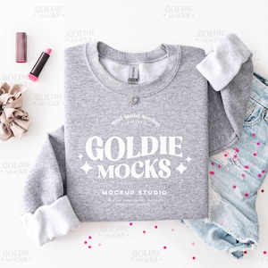 May include: A gray sweatshirt with the text "GOLDIE MOCKS" in white, paired with denim shorts and accessories. The mockup includes a scrunchie, lipstick, and scattered confetti, creating a casual, lifestyle aesthetic.
