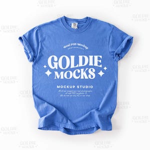 May include: A blue t-shirt with rolled-up sleeves. The shirt features the text "GOLDIE MOCKS" in large white letters, along with the words "Real POD Mockup" and "MOCKUP STUDIO". The shirt is a mockup for POD products.