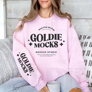 May include: A light pink sweatshirt with the text "GOLDIE MOCKS" in a black, stylized font. The sweatshirt also has the text "MOCKUP STUDIO" and "Real POD Mockup" printed on it. The sleeve has the text "GOLDIE MOCKS" printed on it.