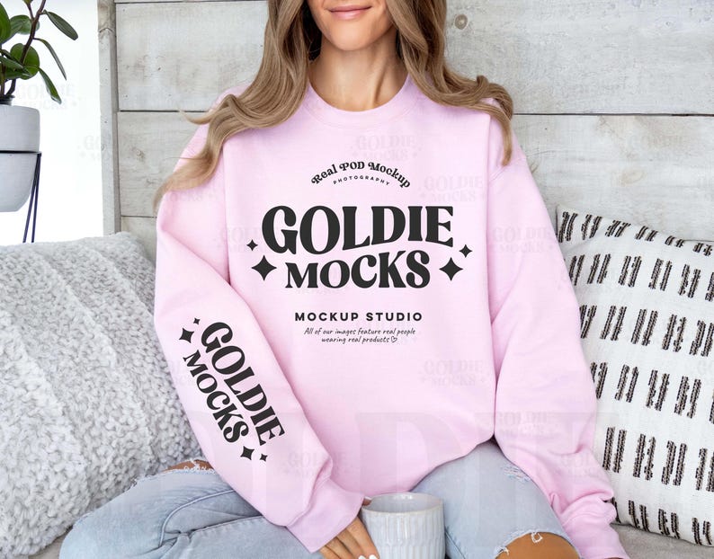 May include: A light pink sweatshirt with the text "GOLDIE MOCKS" in a black, stylized font. The sweatshirt also has the text "MOCKUP STUDIO" and "Real POD Mockup" printed on it. The sleeve has the text "GOLDIE MOCKS" printed on it.