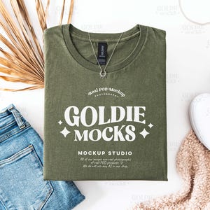 以下が含まれることがあります： 白いグラフィックに「Goldie + Mocks +」と「Mockup Studio」と書かれた緑色のTシャツ。テキストはレトロなフォントスタイルです。Tシャツは白い表面に置かれ、白いスニーカーと茶色のふわふわの毛布が置かれています。