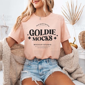 Peut inclure: Une femme portant un t-shirt rose clair avec le texte "Goldie Mocks" en noir. Le t-shirt porte également le texte "Mockup Studio" en noir. La femme porte un short en jean bleu et une veste en fourrure marron clair.