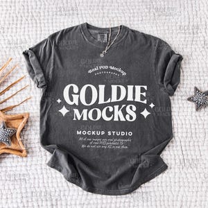 Peut inclure: Un t-shirt gris foncé avec un motif blanc qui dit "Goldie Mocks" et "Real Pod Mockup Photography" avec une étoile blanche de chaque côté du texte. Le t-shirt est posé sur une surface texturée blanche.