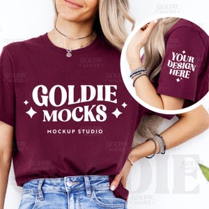May include: A maroon t-shirt with short sleeves featuring the text "Goldie Mocks" in white with a retro font and a diamond design on either side. The text "Mockup Studio" is printed below the main text. The t-shirt is being worn by a person with long blonde hair.