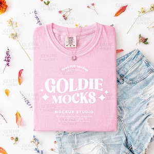 Puede incluir: Una composición plana con una camiseta rosa claro con el texto blanco "GOLDIE MOCKS", combinada con pantalones cortos de mezclilla. La camiseta tiene una etiqueta que dice "COMFORT COLORS". La escena está decorada con flores y hojas coloridas, creando una estética elegante y femenina.