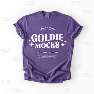 May include: A purple t-shirt with rolled-up sleeves. The shirt features the text "GOLDIE MOCKS" in a white, stylized font, along with the words "MOCKUP STUDIO" and additional text. The shirt is a solid color and appears to be made of soft material.