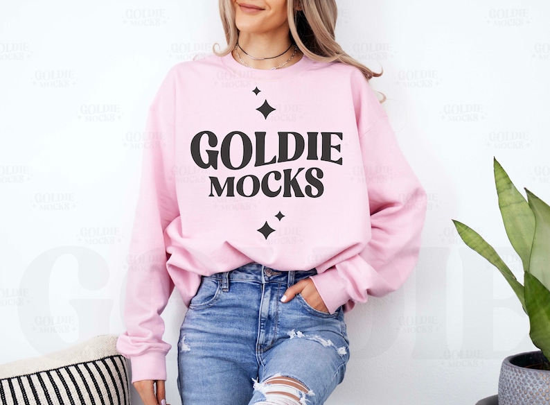 K&ouml;nnte beinhalten: Ein rosa Sweatshirt mit dem Text "GOLDIE MOCKS" in schwarzen Buchstaben. Das Sweatshirt hat einen Rundhalsausschnitt und lange &Auml;rmel. Das Model tr&auml;gt eine blaue Jeans.