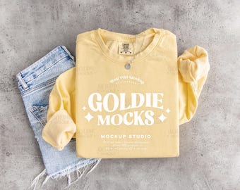 May include: A pale yellow long-sleeved sweatshirt with the text "GOLDIE MOCKS" in white, paired with denim shorts. A silver necklace with a pendant is on the sweatshirt. The sweatshirt has a tag that says "COMFORT COLORS".