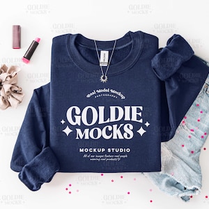 May include: A navy blue sweatshirt with the text "GOLDIE MOCKS" in white, paired with light-wash denim jeans. A necklace with a sun pendant, lipstick, and a scrunchie are also visible. The image is a mockup.