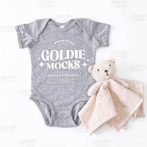 May include: A gray baby onesie with short sleeves and a white graphic that says "Goldie Mocks Mockup Studio". A beige plush toy is lying next to the onesie.