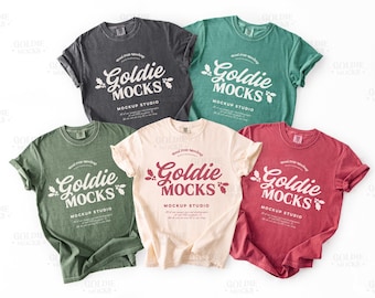 May include: A collection of five t-shirts in various colors: charcoal, teal, olive green, cream, and red. Each shirt features the text "Goldie Mocks Mockup Studio" in a stylized font. The shirts are displayed with rolled-up sleeves.