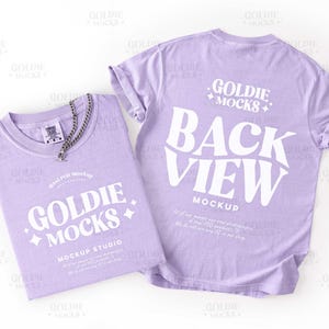 May include: Two lilac t-shirts, one folded with a silver chain necklace, the other unfolded. The folded shirt has the text "GOLDIE MOCKS MOCKUP STUDIO". The unfolded shirt's back says "BACK VIEW".