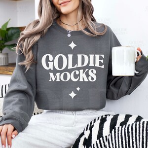 May include: A charcoal grey sweatshirt with the text "Goldie Mocks" in white lettering. The sweatshirt has a relaxed fit and a crew neckline.