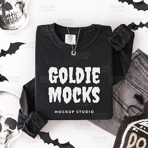 May include: Black long-sleeve shirt with the text "GOLDIE MOCKS" in white. The shirt is laid flat with a pair of gray jeans, a skull, and bat cutouts. A silver necklace with a sun pendant is on the shirt.
