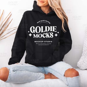 Könnte beinhalten: Ein schwarzer Hoodie mit einem weißen Logo, auf dem "Goldie Mocks" und "Mockup Studio" steht. Der Hoodie wird von einer Person getragen, die auf einer weißen Oberfläche sitzt.