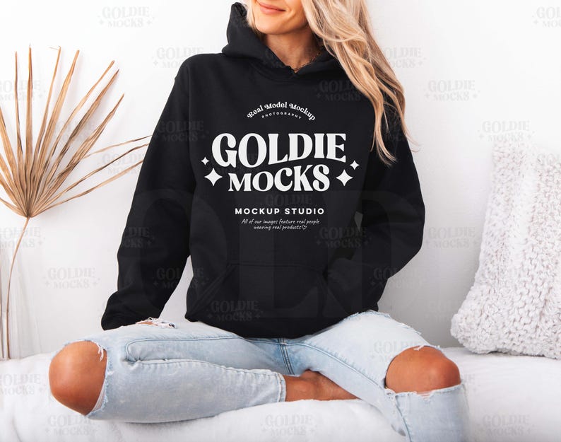 May include: A black hoodie with a white logo that says "Goldie Mocks" and "Mockup Studio". The hoodie is being worn by a person sitting on a white surface.