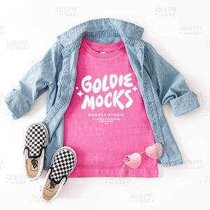 May include: A pink t-shirt with the text "Goldie Mocks Mockup Studio" in white. The t-shirt is laid flat on a white surface with a denim shirt and checkered Vans shoes.