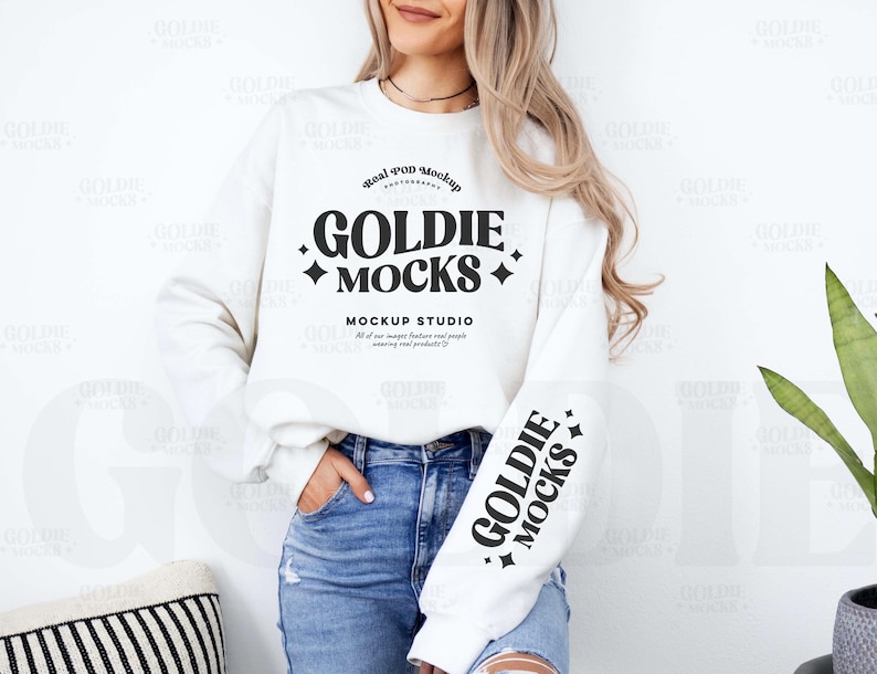 May include: White crewneck sweatshirt with long sleeves and a black graphic print that says "Real POD Mockup Goldie Mocks Mockup Studio". The graphic print features a star design and the text is in a bold, retro font.