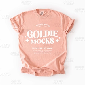 以下が含まれることがあります： 前面に「Goldie Mocks」と「Mockup Studio」の文字が印刷された、ピーチ色のTシャツのモックアップです。