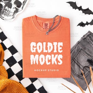 May include: An orange t-shirt with the text "GOLDIE MOCKS" and "MOCKUP STUDIO" in white. The shirt is folded and accessorized with a silver chain necklace, gray jeans, and black bat cutouts. A skull and a black and white checkered pattern are also present.