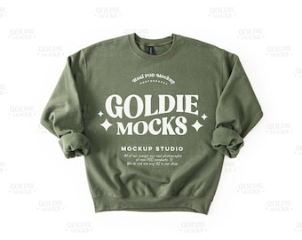 May include: An olive green sweatshirt with the text "GOLDIE MOCKS" in white, along with the words "Real POD Mockup Photography" and "MOCKUP STUDIO". The sweatshirt has a crew neck and rolled-up sleeves.