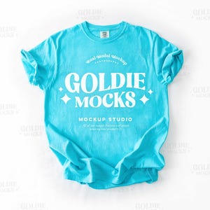 Puede incluir: Una camiseta azul turquesa con un diseño blanco que dice "GOLDIE MOCKS" y "MOCKUP STUDIO".