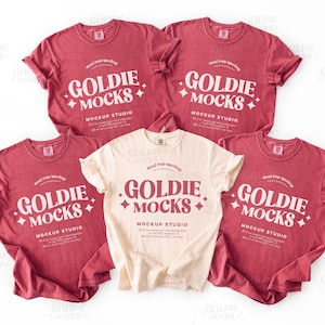 May include: Five t-shirts in a mockup studio setting. Four are a reddish-pink colour, and one is cream-coloured. Each shirt has the text "GOLDIE MOCKS" in a white, stylised font, with the words "MOCKUP STUDIO" below.