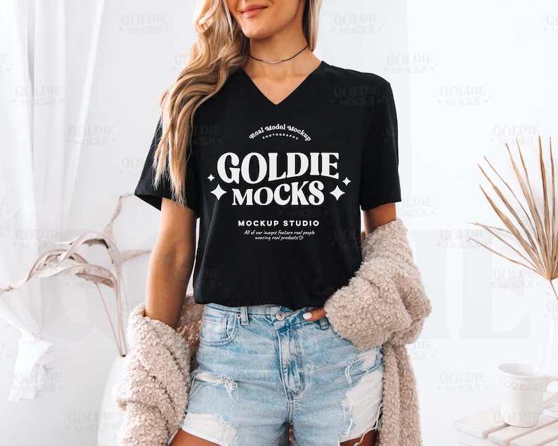 May include: A black v-neck t-shirt with the text "GOLDIE MOCKS MOCKUP STUDIO" in white. The text is in a retro font style with stars on either side. The shirt is being worn by a person with long brown hair.