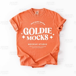 Puede incluir: Una camiseta naranja con las mangas enrolladas. La camiseta presenta el texto "GOLDIE MOCKS" en una fuente blanca estilizada, junto con las palabras "MOCKUP STUDIO" y otro texto. La camiseta es una maqueta de modelo real.