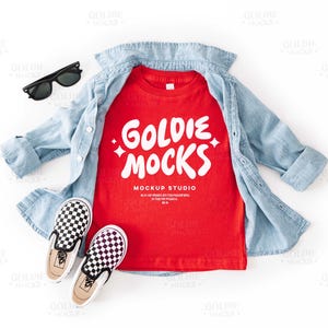 Puede incluir: Camiseta roja con el texto "Goldie Mocks Mockup Studio" en letras blancas. La camiseta está colocada sobre una superficie blanca con una camisa vaquera azul y zapatos de tela negros y blancos a cuadros.