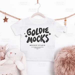 May include: A white t-shirt with the text "GOLDIE MOCKS MOCKUP STUDIO" in black, hanging on a wooden hanger. The shirt is surrounded by decorative elements, including a star, beads, and a fluffy pink cushion.