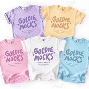 Op de afbeelding: Vijf pastelkleurige t-shirts worden getoond, elk met de tekst "GOLDIE MOCKS" in een speels lettertype. De shirts zijn in tinten lavendel, geel, roze, wit en lichtblauw. De woorden "MOCKUP STUDIO" staan eronder.