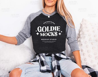 May include: A charcoal gray and heather gray raglan sleeve t-shirt with the text "GOLDIE MOCKS" in white. The shirt is part of a mockup studio collection. The model is wearing light blue jeans and a black and white plaid shirt.