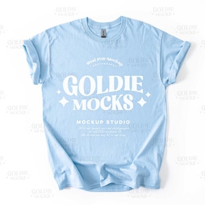 May include: A light blue t-shirt with the text "Goldie Mocks" in white. The text is in a retro font style and is surrounded by white stars. The text "Real POD Mockup" and "Mockup Studio" are also visible in white.