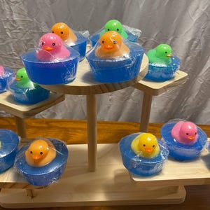 Op de afbeelding: Een houten displaystandaard bevat verschillende kleurrijke eendvormige zepen. De zepen zitten in blauwe bakjes en zijn verpakt in doorzichtig plastic. De eenden zijn geel, oranje, roze en groen.